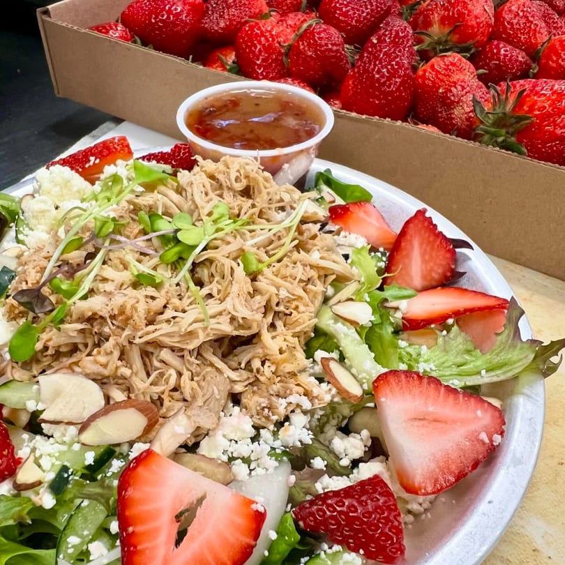 A Farm Fresh Fast salad with shredded chicken, strawberries, feta, almonds, and dressing, next to a box of strawberries.